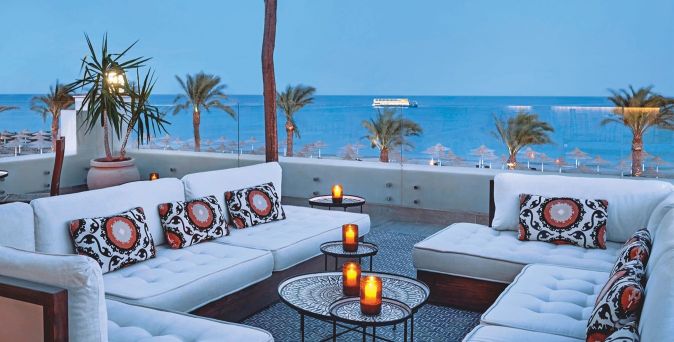 Lounge-Bereich mit weißen Sofas und bunten Kissen, Blick auf das Meer und Palmen, mehrere Kerzen auf Tischen.