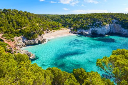 Panorama der Bucht Cala Mitjaneta auf Menorca mit feinem weißen Sand, türkisfarbenem Meer und dichter grüner Vegetation.