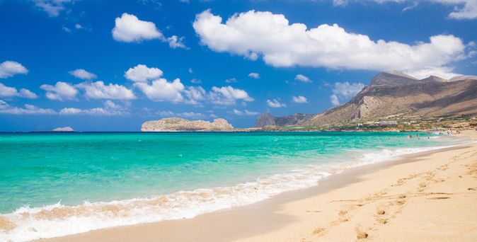 Sandstrand mit türkisfarbenem Meer und bewölktem Himmel, im Hintergrund Berge und Küstenlinie auf Kreta