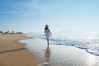 Frau in weißem Kleid und Strohhut spaziert barfuß am Strand entlang, während sanfte Wellen den Sand berühren