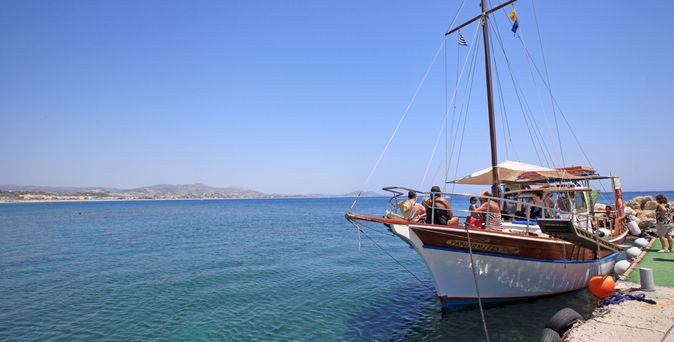 Mehrere Personen sitzen auf einem hölzernen Segelboot, das an einem Steg im Meer vor einer Küstenlandschaft festgemacht ist.