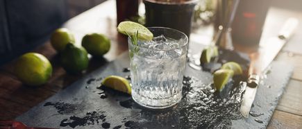 Ein Glas Gin mit Eiswürfeln und Limettenscheibe steht auf einem Tisch, daneben liegen frische Limetten