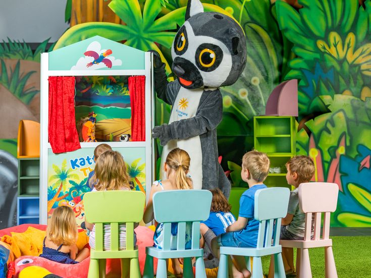 Katta-Maskottchen spielt ein buntes Puppentheater vor sitzenden Kindern in einem liebevoll gestalteten Indoor-Spielbereich mit tropischer Kulisse