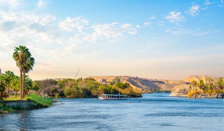 Ein Ausflugsboot fährt über den Nil in Ägypten, umgeben von Palmen, Felsformationen und sanften Wüstenhügeln unter einem klaren Himmel mit vereinzelten Wolken