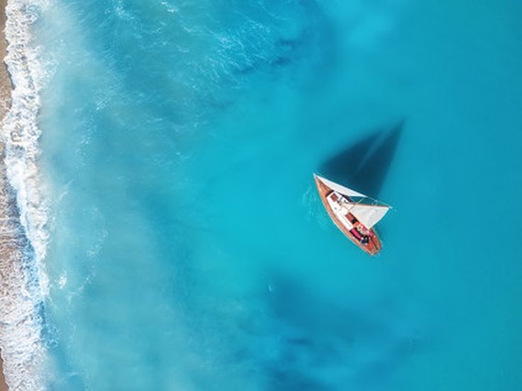 Luftaufnahme eines Segelboots auf türkisblauem Meer nahe eines Sandstrandes.
