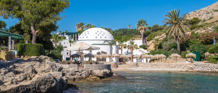 Küstenbereich mit Felsen, klarem Wasser, weißen Gebäuden mit Kuppeldach und mehreren Sonnenschirmen, umgeben von Palmen und Pinien auf Rhodos.