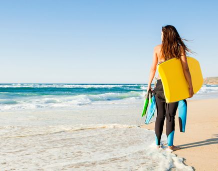 Frau im Neoprenanzug steht mit Bodyboard und Flossen am Strand und blickt auf das Meer mit Wellen