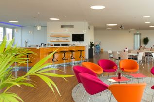 Staffelgeschoss in Gebäude 2 von schauinsland-reisen mit moderner Lounge- und Barzone, orangefarbener Theke mit Barhockern, farbigen Sesseln, Tischen und heller, offener Raumgestaltung