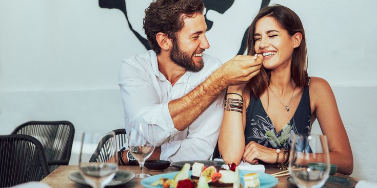 Ein Mann füttert lachend eine Frau in einem Restaurant, beide sitzen an einem Tisch mit Dessert und Weingläsern