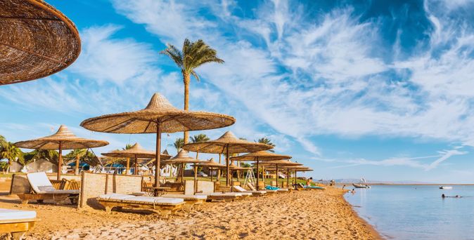 Strand am Roten Meer in Ägypten mit strohbedeckten Sonnenschirmen, Liegen und ruhigem, flach abfallendem Wasser unter blauem Himmel