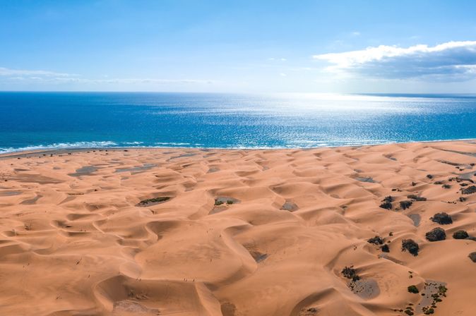 Sanddünen im Vordergrund mit Spuren, dahinter blauer Ozean unter klarem Himmel