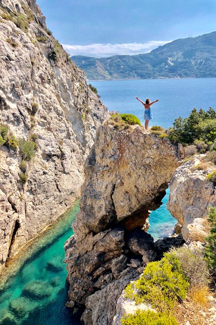 Person in blauem Kleid steht mit erhobenen Armen auf einem Felsen über türkisfarbenem Wasser an der Küste von Korfu.