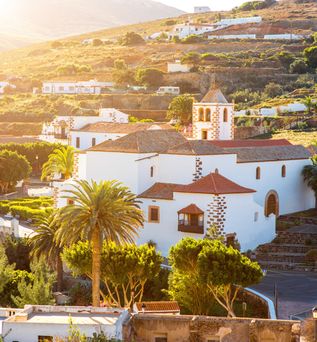 Weiß getünchte Gebäude mit roten Dächern und einem Kirchturm in Betancuria, Fuerteventura, umgeben von Palmen und hügeliger Landschaft im Sonnenlicht.