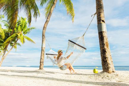 Person sitzt in einer Hängematte zwischen Palmen an einem weißen tropischen Sandstrand mit Blick auf das ruhige Meer