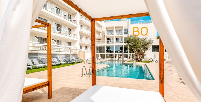Blick von einer überdachten Liege auf einen rechteckigen Pool mit Holzumrandung und Liegestühlen vor einem mehrstöckigen Hotelgebäude mit Balkonen und dem Schriftzug 'BQ'