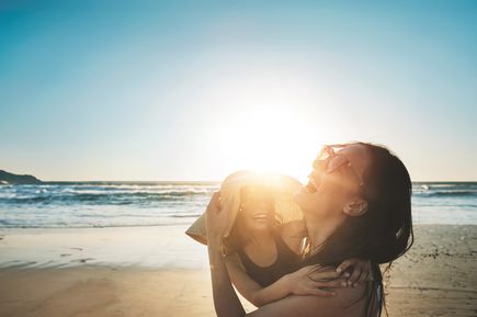 Frau hält ein lachendes Kind am Strand im warmen Gegenlicht der untergehenden Sonne, während im Hintergrund sanfte Wellen ans Ufer rollen