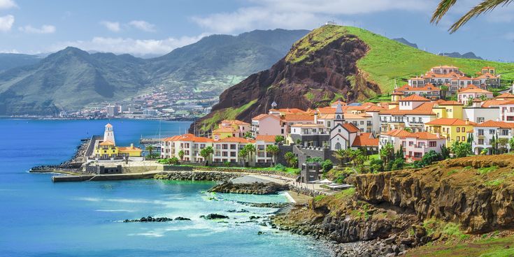 Blick auf das Küstendorf Quinta do Lorde auf Madeira mit Häusern im portugiesischen Stil, kleinem Leuchtturm, felsiger Küste, ruhiger Bucht und grünen Bergen im Hintergrund