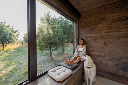 Person sitzt auf einer Fensterbank in einem Holzraum neben einem Hund, mit Blick durch eine große Glasfront auf eine grüne Landschaft mit Bäumen.