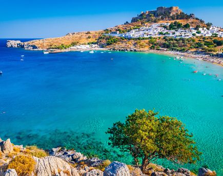 Bucht mit türkisfarbenem Wasser, Segelboot, felsigem Ufer und weißem Dorf unter einer Burg auf einem Hügel.