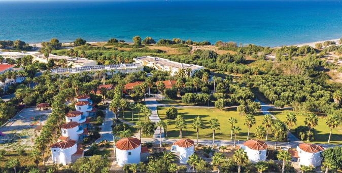 Luftaufnahme des Eurovillage Achilleas Hotels auf Kos mit mehreren weißen Windmühlen, grünen Gärten und Blick auf das Meer im Hintergrund