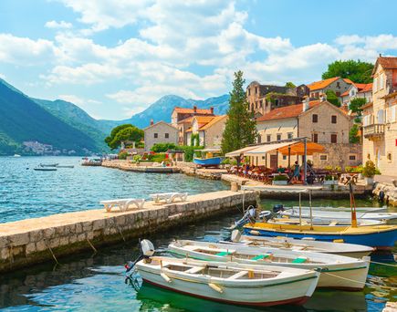 Kleiner Hafen mit mehreren kleinen Booten am Ufer eines klaren Gewässers, umgeben von Häusern mit roten Dächern und Bergen im Hintergrund