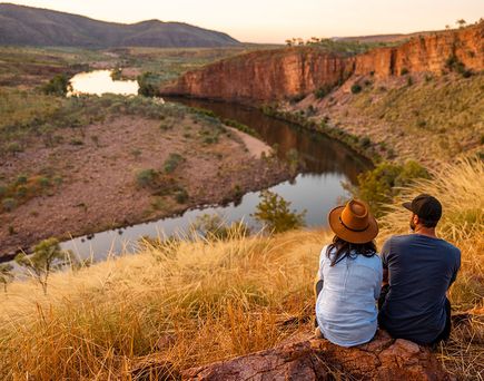 Zwei Personen sitzen auf einem Felsen und blicken auf einen Fluss, der sich durch eine trockene, hügelige Landschaft schlängelt.