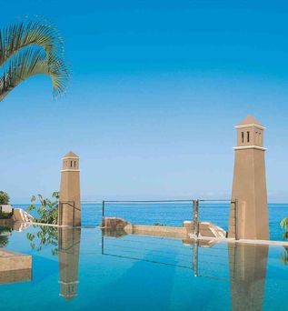 Zwei braune Türme neben einem Infinity-Pool mit Blick auf das Meer und einen klaren blauen Himmel, links eine Palme.