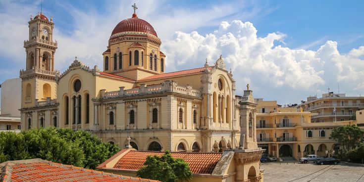 Große Kirche mit roter Kuppel und Glockenturm vor bewölktem Himmel, umgeben von Bäumen und Gebäuden.