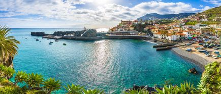 Panoramablick auf den Küstenort Câmara de Lobos auf Madeira mit türkisblauer Bucht, Fischerbooten im Hafen, bunten Häusern an den Berghängen und subtropischer Vegetation im Vordergrund