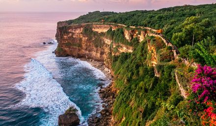 Steile Uluwatu‑Klippe auf Bali in Indonesien mit üppiger grüner Vegetation, einem Weg entlang der Felskante und Wellen, die gegen den Fuß der Klippen schlagen