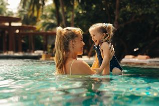 Frau hebt ein kleines Kind spielerisch im warm beleuchteten Pool hoch, umgeben von tropischen Pflanzen und Holzterrassen