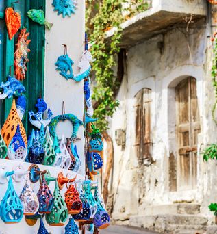 Bunte Keramikfiguren in verschiedenen Formen hängen an einer weißen Wand neben einem engen, sonnigen Gassenabschnitt mit blauen Türen und grünen Pflanzen.