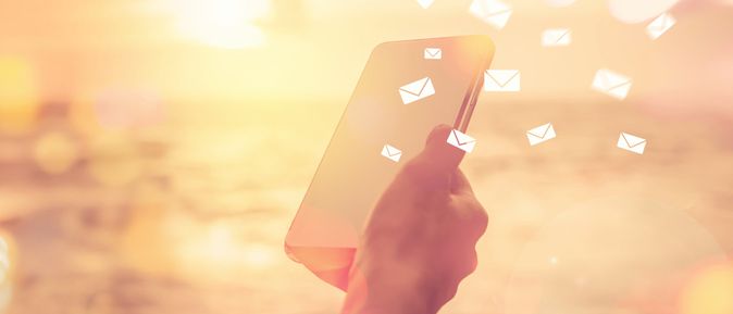 Eine Hand hält ein Smartphone, während kleine, weiße Briefumschläge in der Luft schweben. Im Hintergrund ist eine verschwommene Küstenlandschaft bei Sonnenuntergang zu sehen.