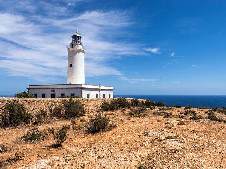 Leuchtturm Far de la Mola auf Formentera mit weißem Turm auf felsiger Küste, niedrigem Gebäudekomplex, karger Vegetation und blauem Meer unter klarem Himmel