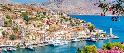 Blick auf die Küstenstadt Symi mit bunten Häusern am Hang, mehreren Booten im Hafen und Bergen im Hintergrund.