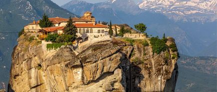 Kloster auf einem hohen Felsen mit roten Dächern, umgeben von Bergen mit schneebedeckten Gipfeln im Hintergrund.