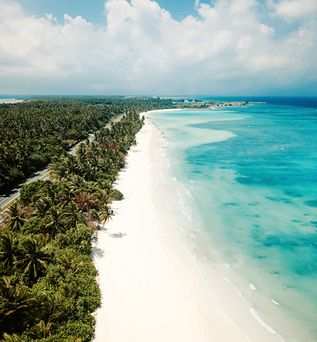 Luftaufnahme von Laamu Gan auf den Malediven mit weißem Sandstrand, türkisfarbenem Indischen Ozean, dichter Palmenvegetation und Küstenstraße entlang der tropischen Insel