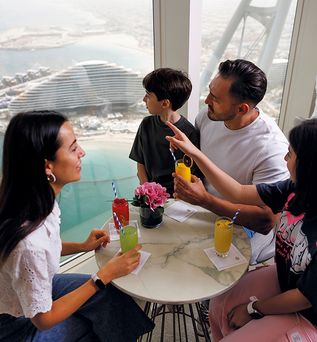 Vier Personen sitzen an einem runden Tisch im Burj Al Aarab mit bunten Getränken und einer kleinen Blumenpflanze, im Hintergrund große Fenster mit Blick auf das Meer und das moderne Gebäude des Jumeirah Marsa Al Arab