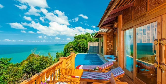 Blick von einer Holzterrasse mit Liegestühlen und Infinity-Pool auf das Meer und bewachsenen Hügel unter blauem Himmel mit Wolken