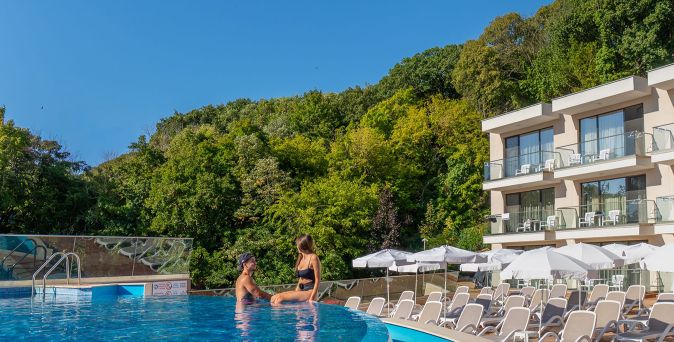 Zwei Personen sitzen am Rand eines Außenpools vor einem Hotelgebäude mit mehreren Balkonen und Liegestühlen unter Sonnenschirmen, umgeben von grünen Bäumen.