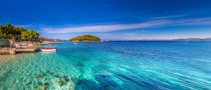 Klares türkisfarbenes Meer mit kleinen Booten am Ufer und bewaldeter Insel im Hintergrund unter blauem Himmel