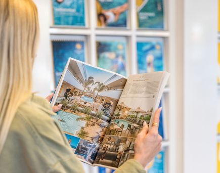 Person mit langen blonden Haaren liest einen Katalog mit Hotelbildern in einem hellen Raum mit Wanddekoration.