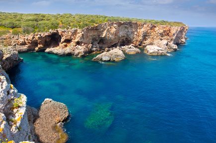 Steile Klippen und felsige Küste von S’Algar auf Menorca, mit tiefblauem Meer und grüner Vegetation im Hintergrund