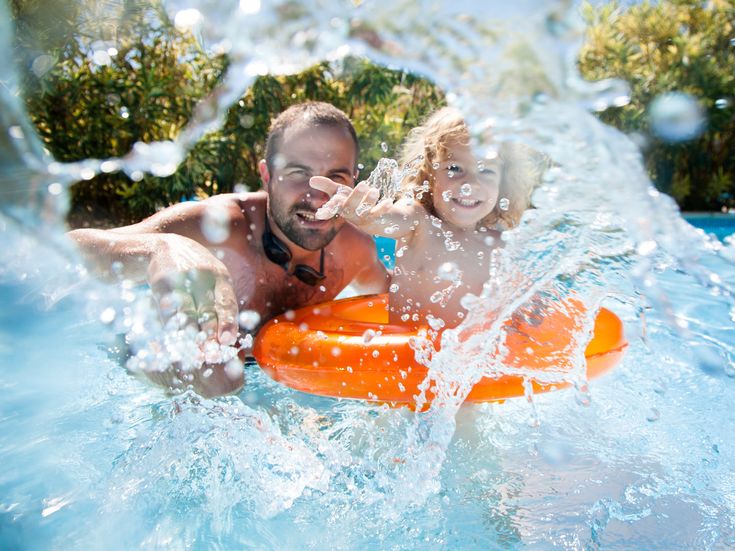 Vater mit Kind im Schwimmreifen, lachend und umgeben von Wasserspritzern in einem Pool