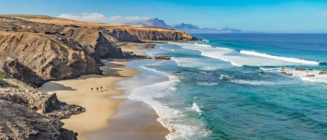 Langer Sandstrand mit Felsen und Klippen links, drei Personen am Strand, rechts türkisfarbenes Meer mit Wellen, blauer Himmel mit Wolken.
