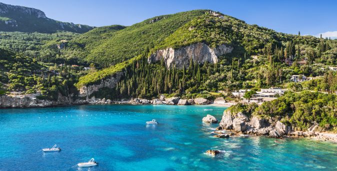 Bucht mit klarem, blauem Wasser, umgeben von grünen Hügeln und Felsen, mehrere Boote auf dem Wasser, Küstenlinie mit Vegetation und Gebäuden in Paleokastritsa auf Korfu.