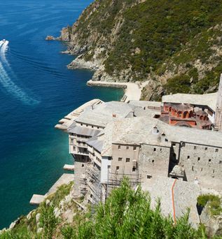 Blick von oben auf ein großes Klostergebäude an einer steilen Küste, davor ein Ausflugsboot auf blauem Wasser mit sichtbarer Bugwelle.