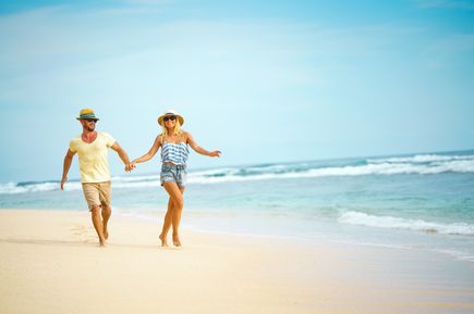 Ein lachendes Paar läuft Hand in Hand am Strand entlang, bei sonnigem Wetter und leichtem Wellengang