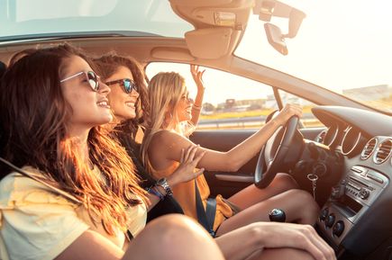Drei junge Frauen sitzen im Auto, eine fährt, während die anderen lächelnd aus dem Fenster schauen. Die warme Abendsonne scheint ins Fahrzeug und sorgt für eine entspannte, sommerliche Stimmung während der Fahrt