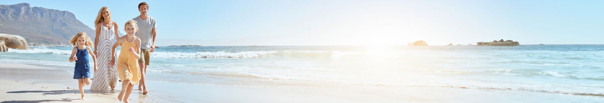 Familie mit zwei Kindern läuft lachend am Strand entlang, im Hintergrund Meer und Berge bei strahlend blauem Himmel